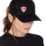 Front view of a female wearing a black classic trucker hat with a pink embroidered double diamond shield on the bottom right panel