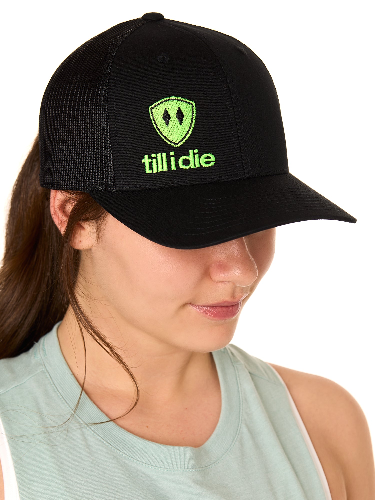 Front view of a female model wearing a black classic trucker hat, featuring a lime green embroidered Till I Die logo on the lower right front panel.