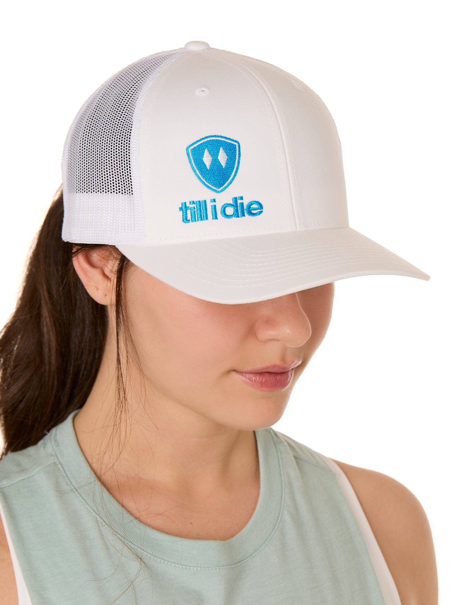 Front view of a female model wearing a white classic trucker hat, featuring a blue embroidered Till I Die logo on the lower right front panel.