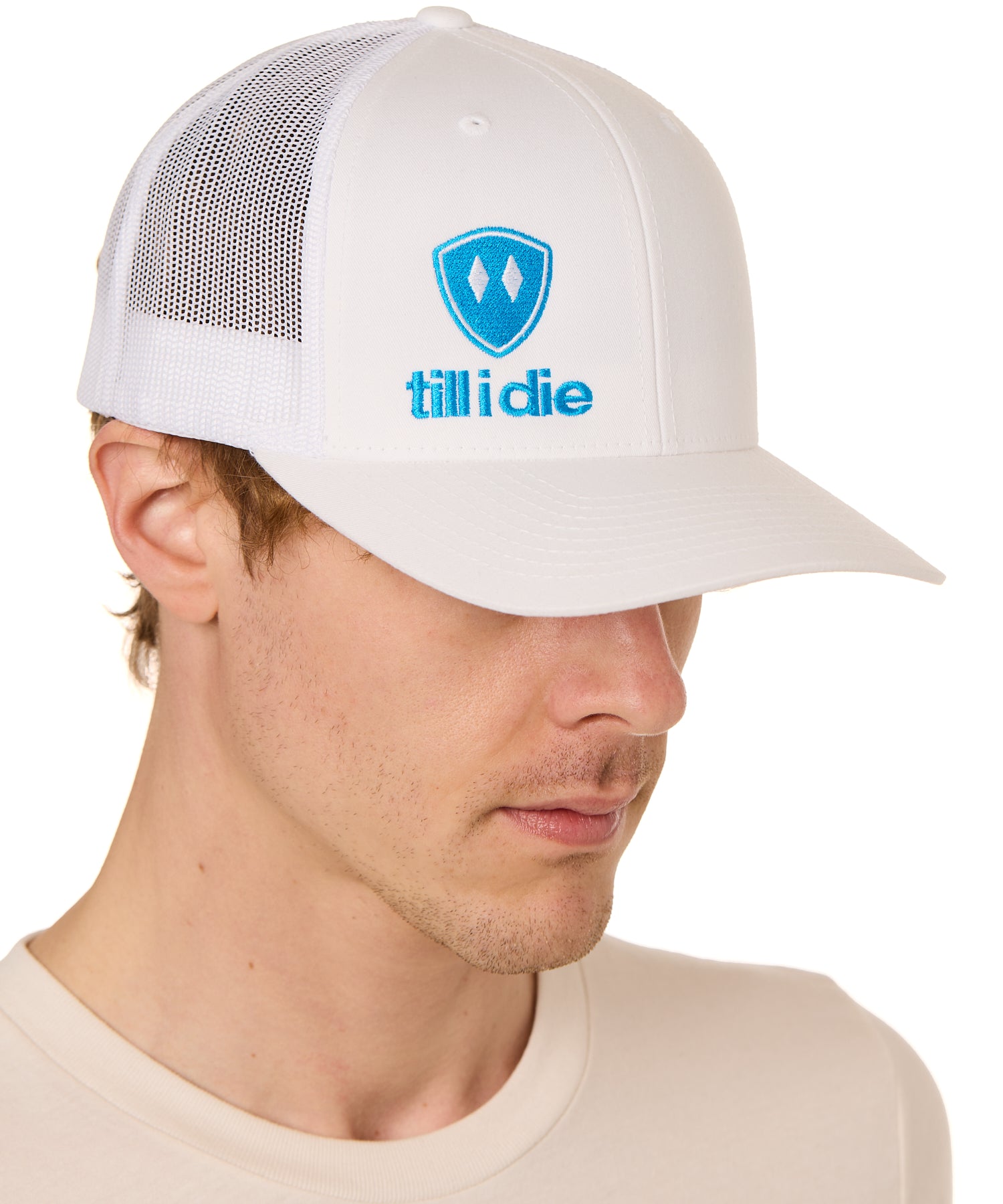 Front view of a male model wearing a white classic trucker hat, featuring a blue embroidered Till I Die logo on the lower right front panel.