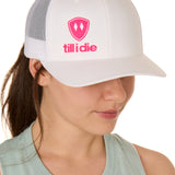 Front view of a female model wearing a white classic trucker hat, featuring a hot pink embroidered Till I Die logo on the lower right front panel.