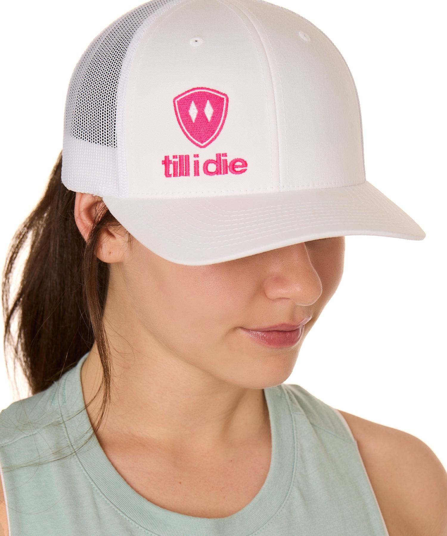 Front view of a female model wearing a white classic trucker hat, featuring a hot pink embroidered Till I Die logo on the lower right front panel.