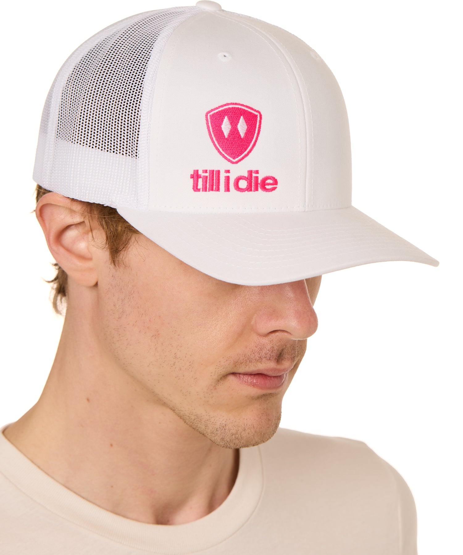 Front view of a male model wearing a white classic trucker hat, featuring a hot pink embroidered Till I Die logo on the lower right front panel.