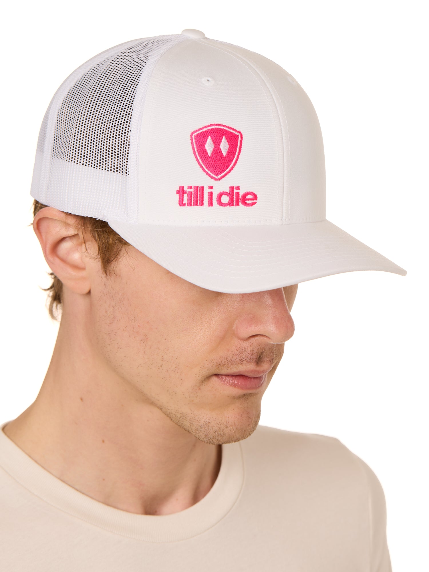 Front view of a male model wearing a white classic trucker hat, featuring a hot pink embroidered Till I Die logo on the lower right front panel.