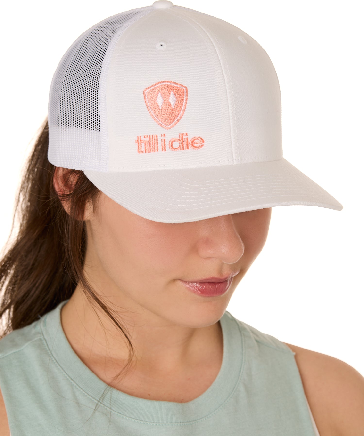 Front view of a female model wearing a white classic trucker hat, featuring a coral embroidered Till I Die logo on the lower right front panel.