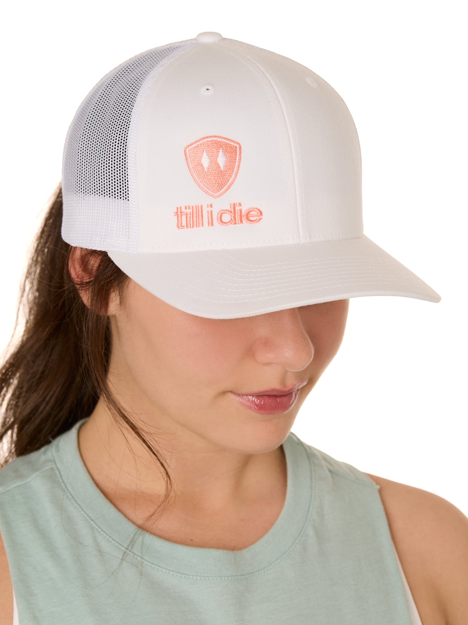 Front view of a female model wearing a white classic trucker hat, featuring a coral embroidered Till I Die logo on the lower right front panel.