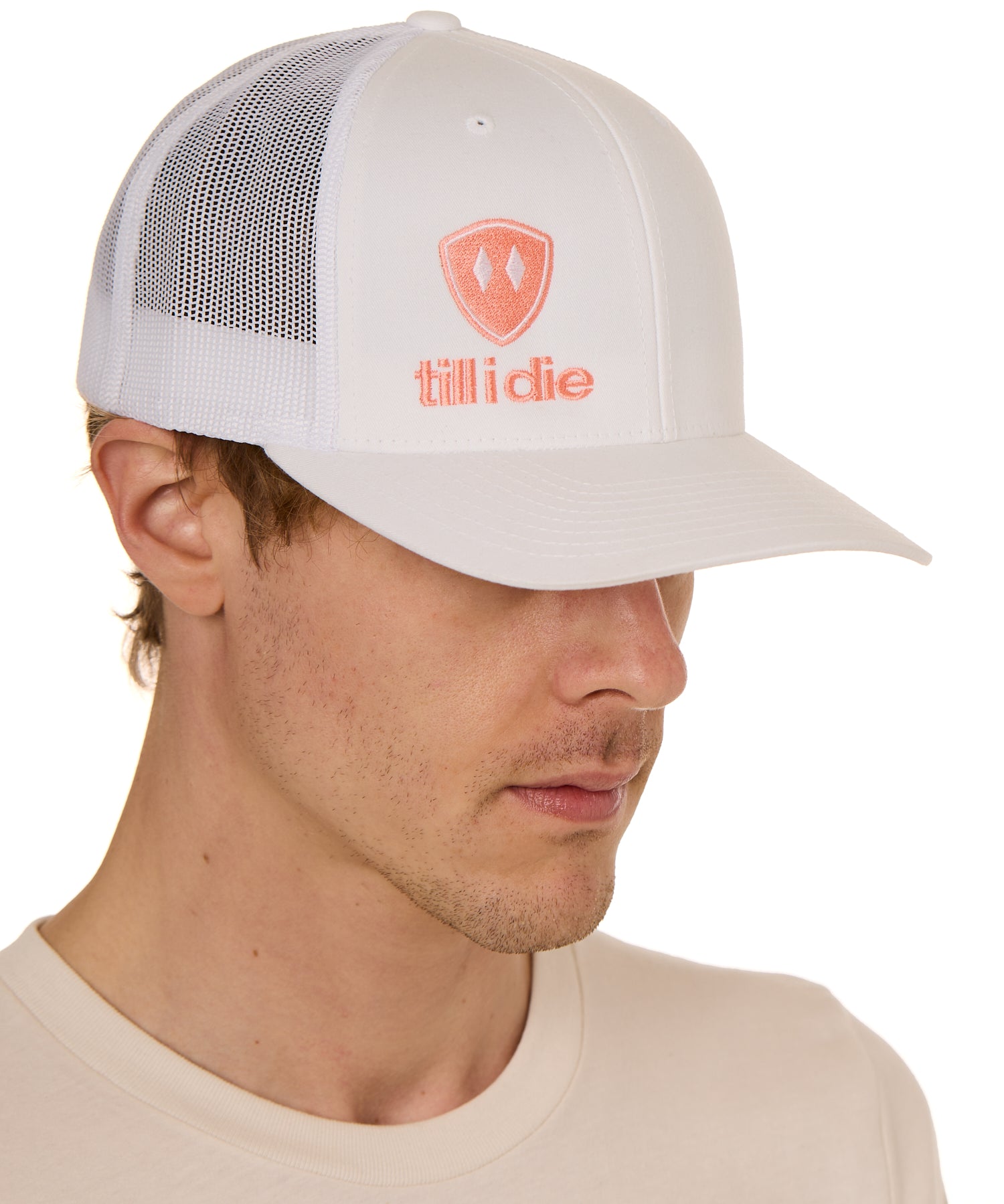 Front view of a male model wearing a white classic trucker hat, featuring a coral embroidered Till I Die logo on the lower right front panel.