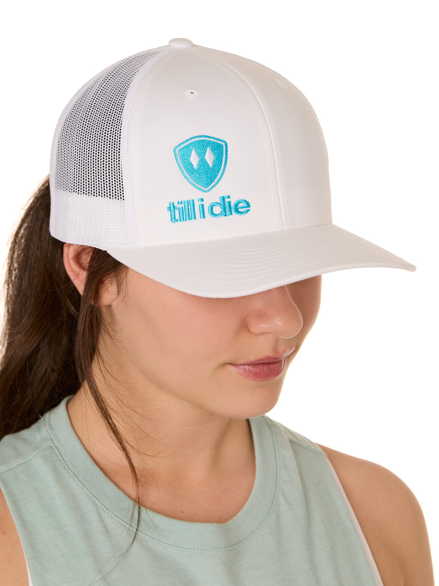 Front view of a female model wearing a white classic trucker hat, featuring a teal embroidered Till I Die logo on the lower right front panel.