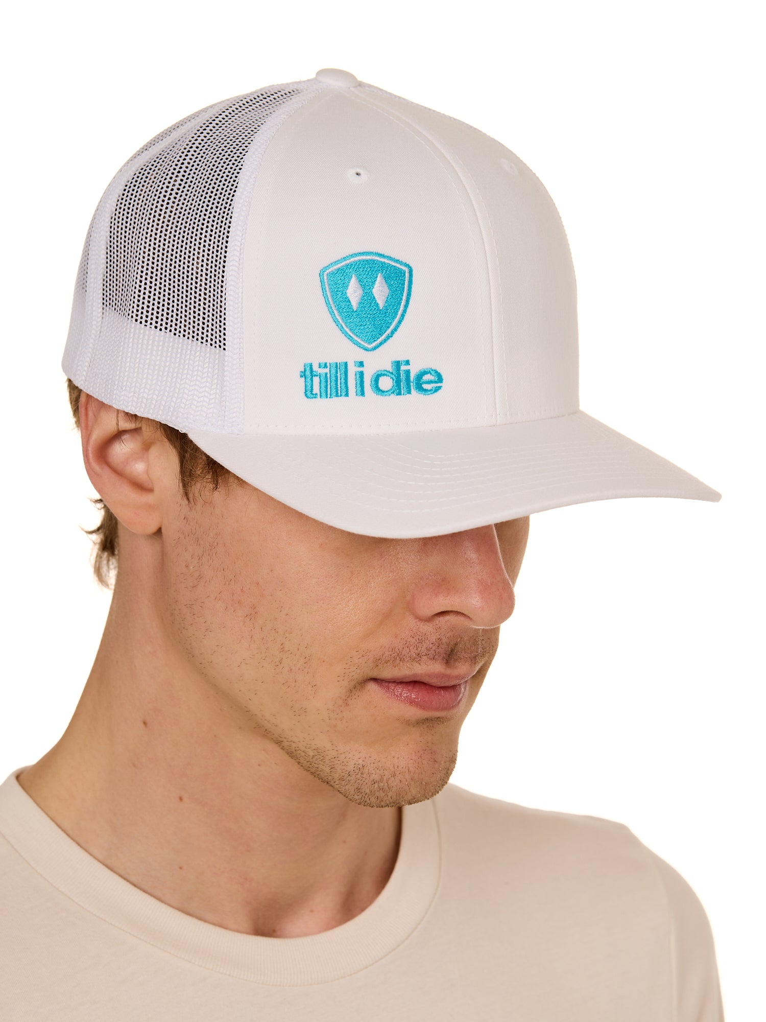 Front view of a male model wearing a white classic trucker hat, featuring a teal embroidered Till I Die logo on the lower right front panel.