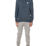 Front view of a male model wearing the Summer night hoodie in washed denim on a white background. The hoodie is embroider with a gray and white  double diamond shield over the wearers left chest. Paired with The Summer Nights mens joggers.