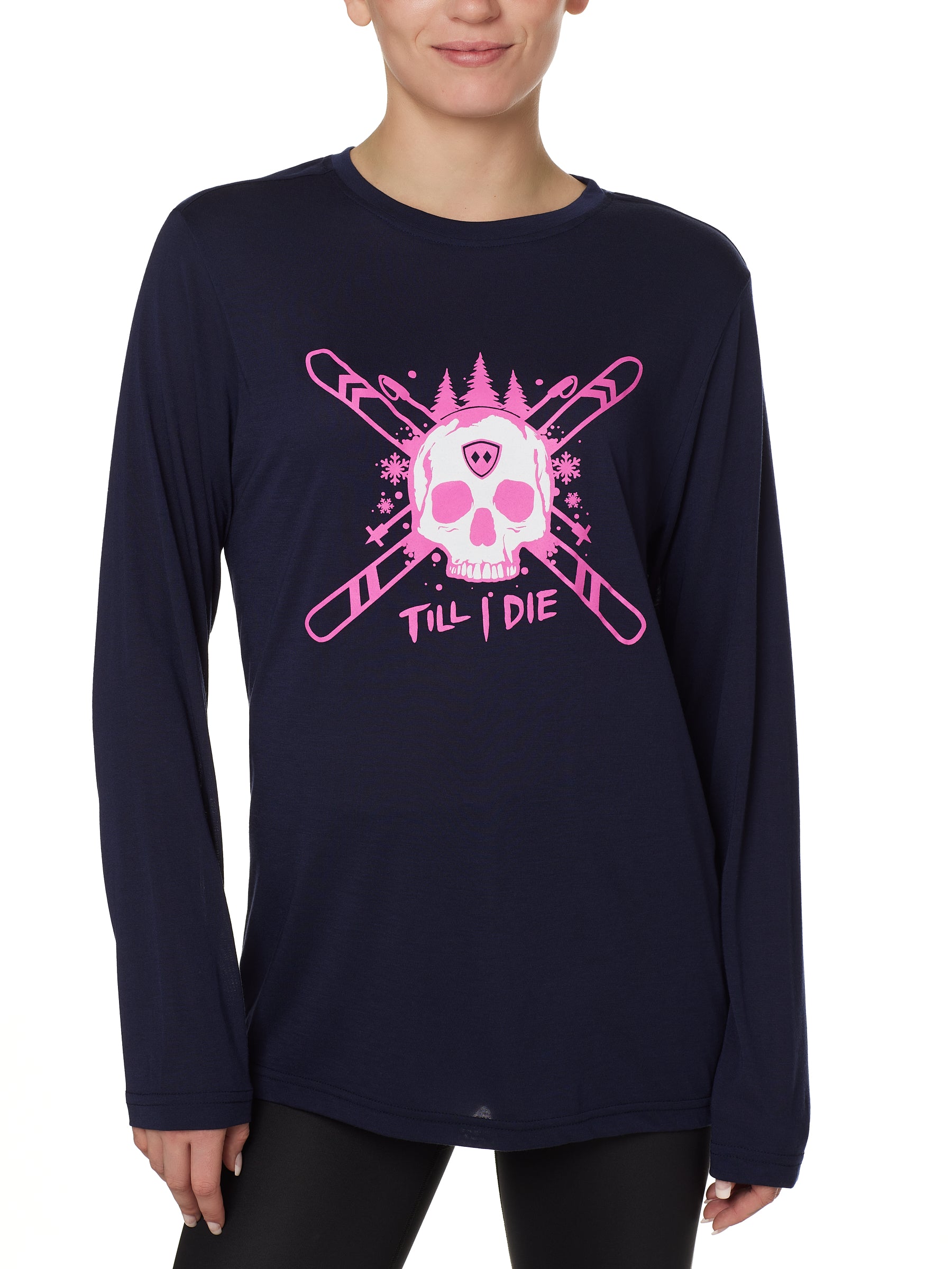 Front view of a female model wearing the ULLR navy crew neck long sleeve tee on a white background. The pink and white graphic features a skull over crossed skis, with snowflakes, pine trees, and “Till I Die” text layered below.
