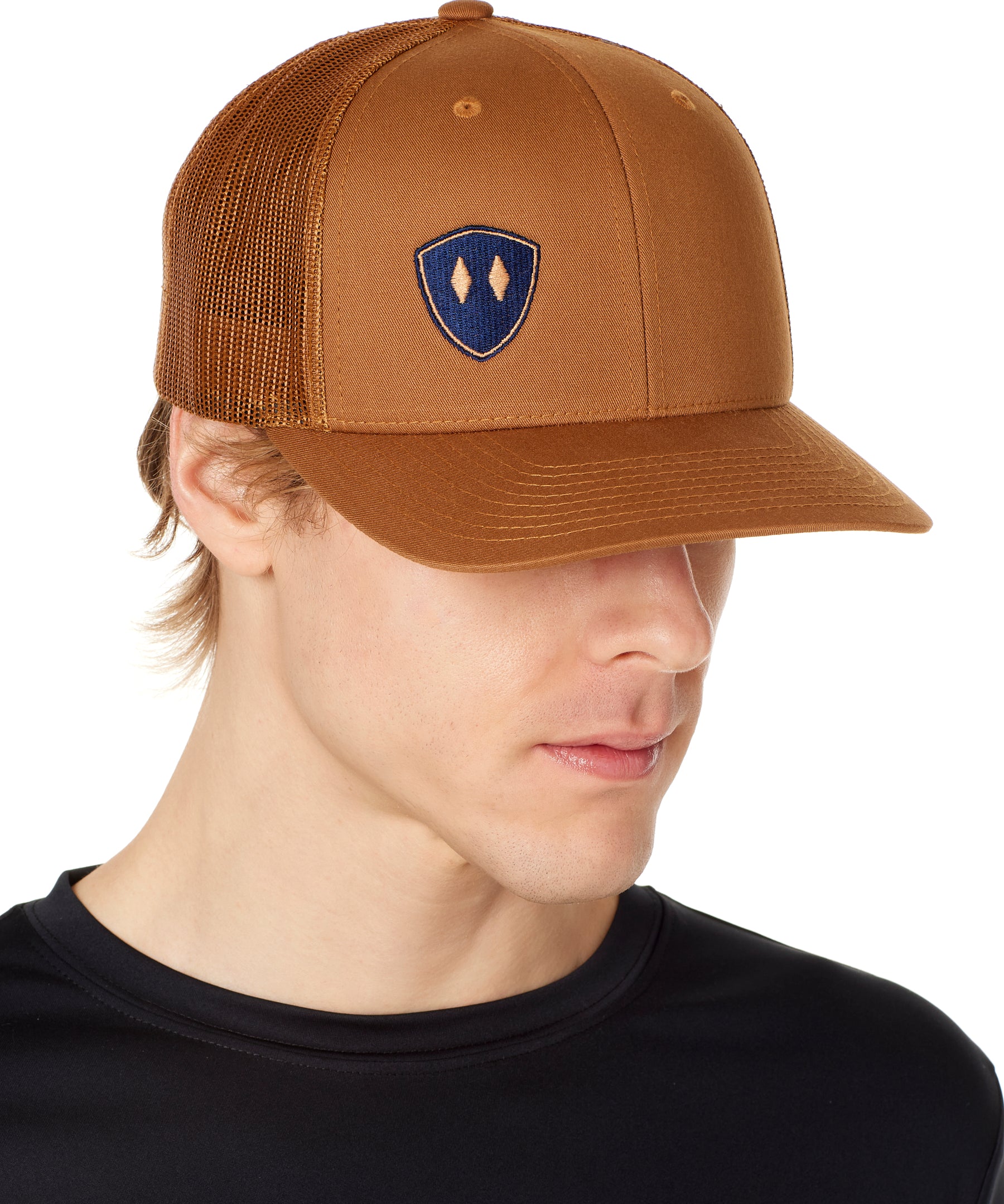 A man wearing a caramel and navy trucker hat with a shield logo on the front.