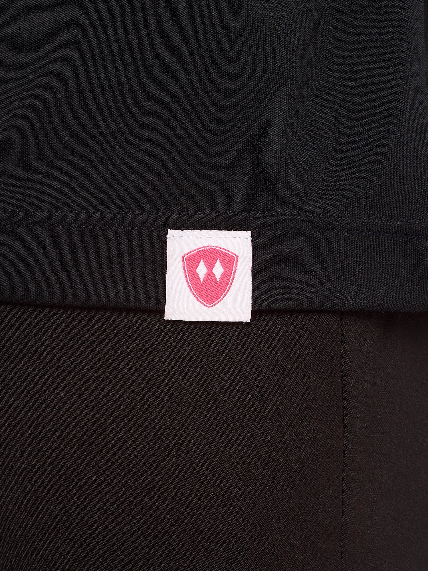 Close-up detail of a pink Double Diamond Shield woven hem label on a black performance long sleeve shirt.