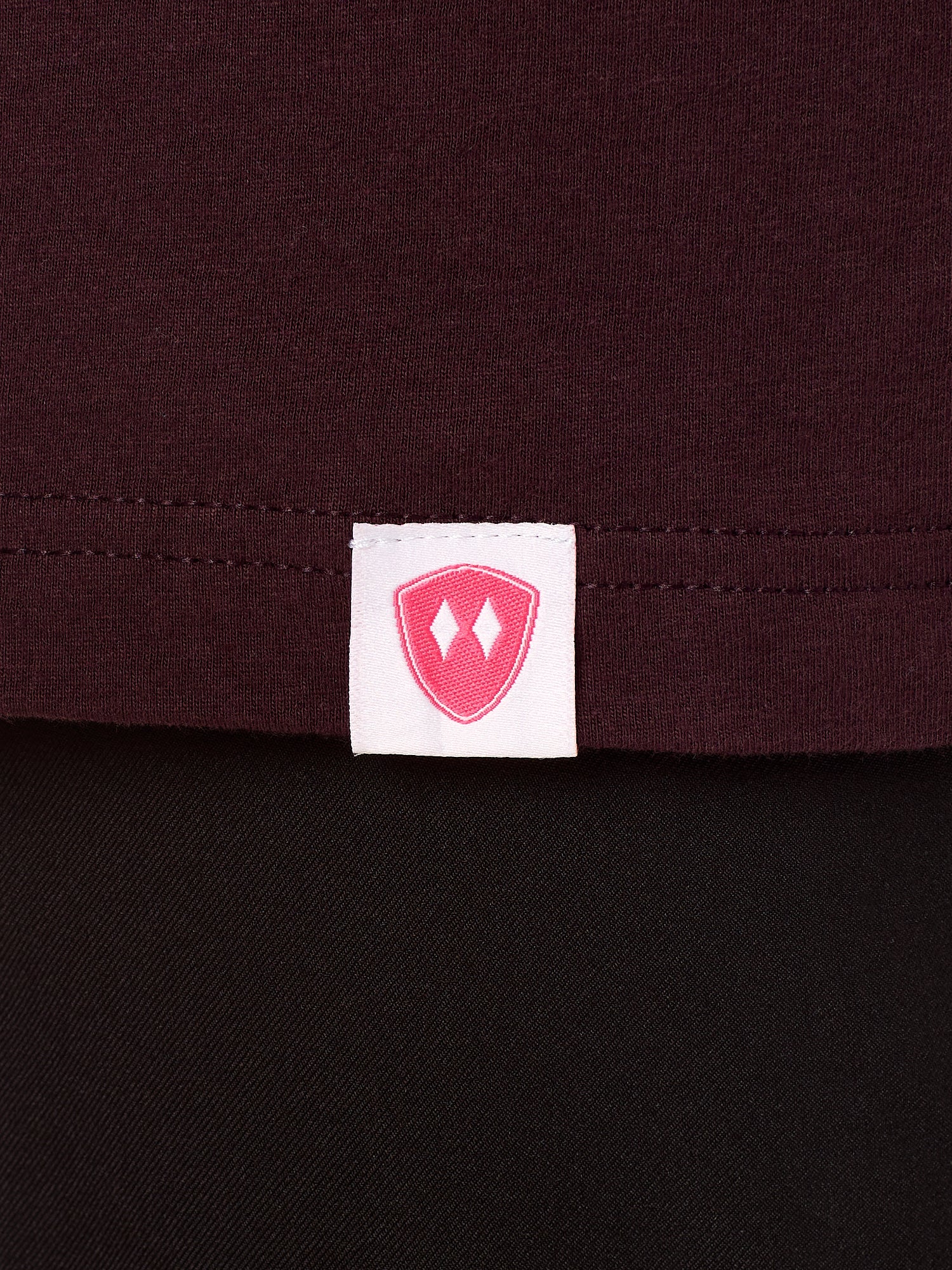 Close-up detail of a pink Double Diamond Shield woven hem label on a Oxblood tee shirt.
