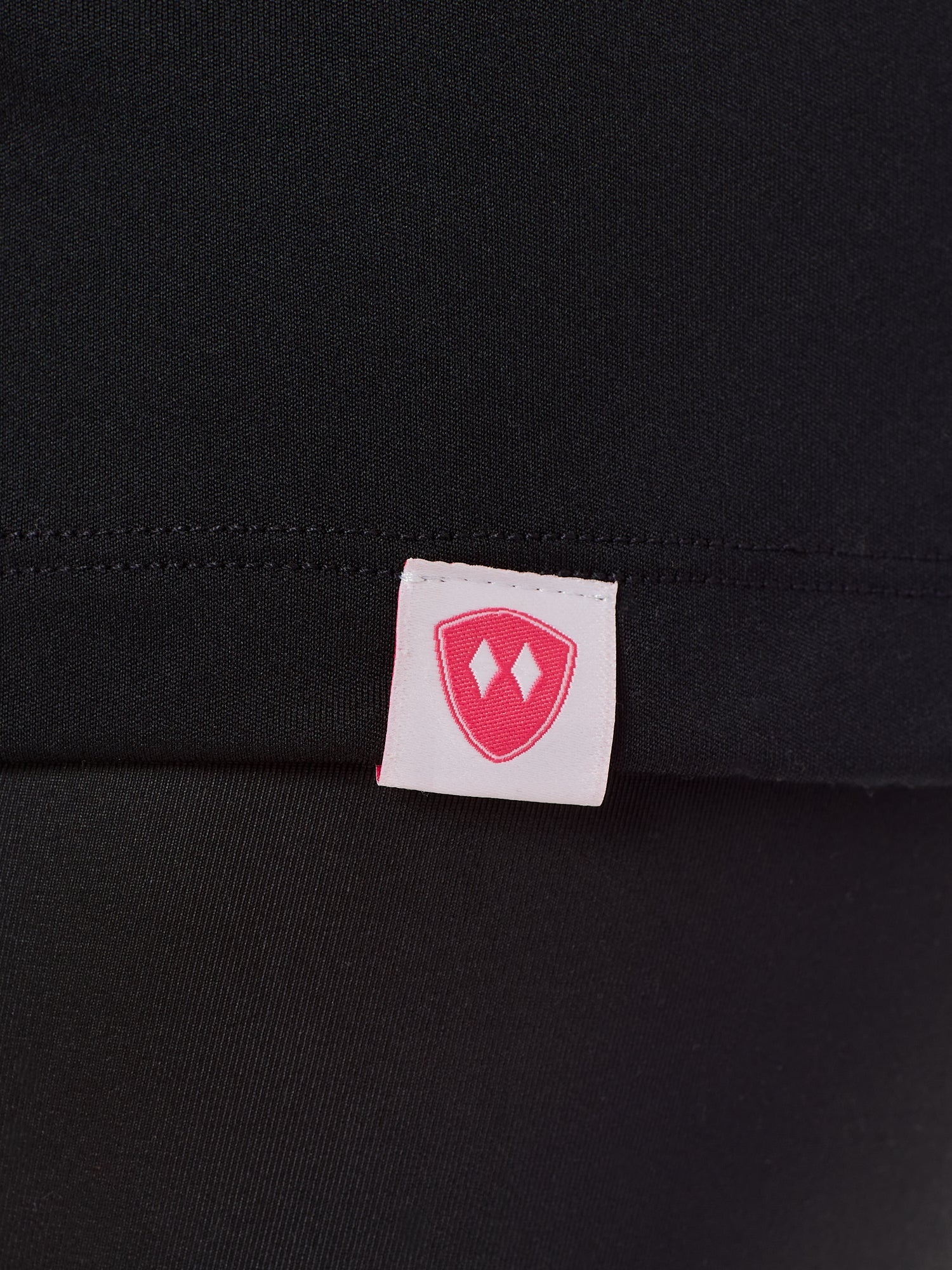 Close-up detail of a pink Double Diamond Shield woven hem label on a black performance long sleeve shirt.