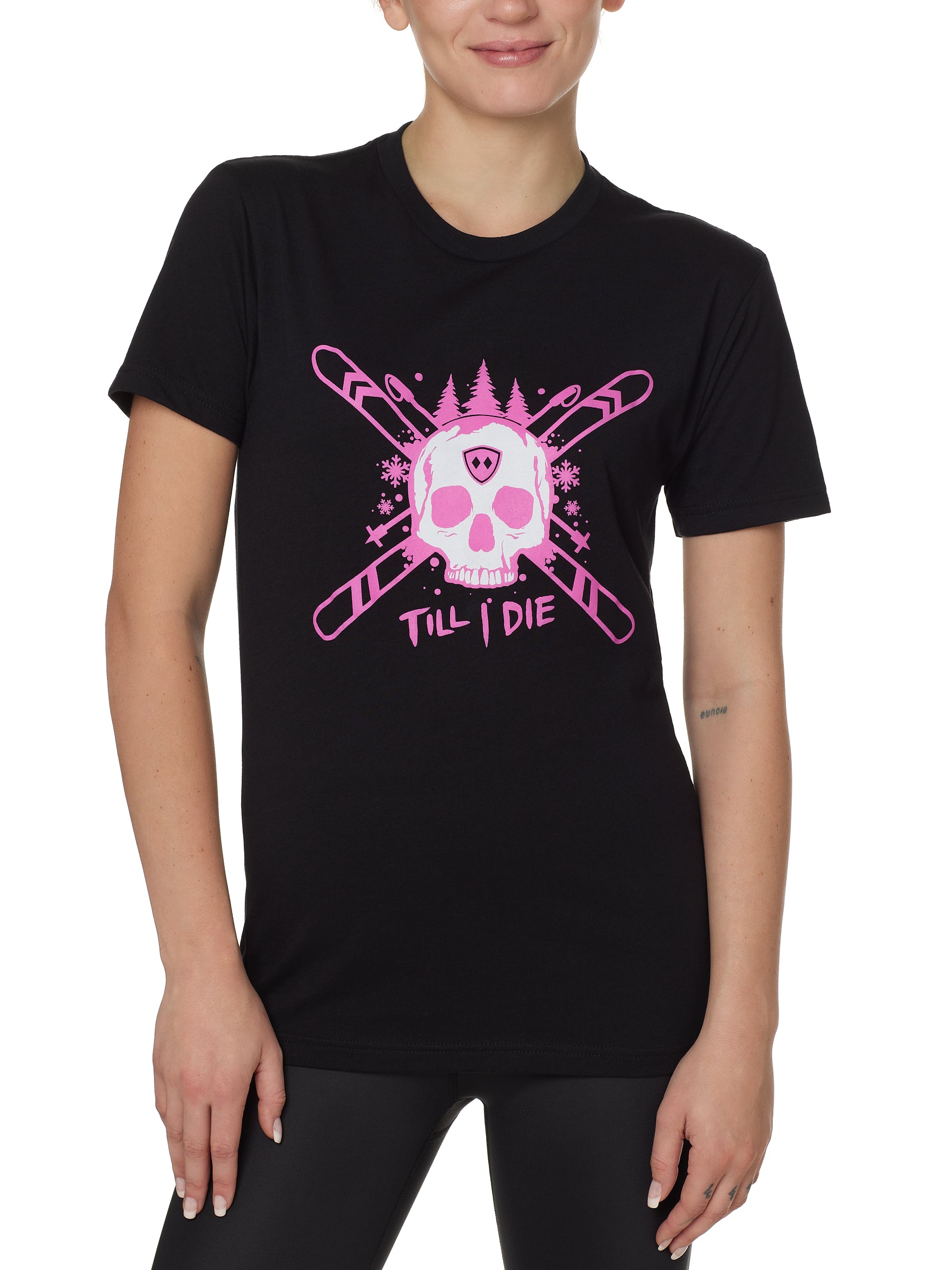 Front view of a female model wearing the ULLR black crew neck tee on a white background. The pink and white graphic features a skull over crossed skis, with snowflakes, pine trees, and “Till I Die” text layered below.