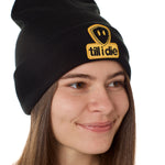 Sideview of a female model wearing a black knit beanie, featuring a cuff and a chenille patch of the Till I Die logo in yellow and black.
