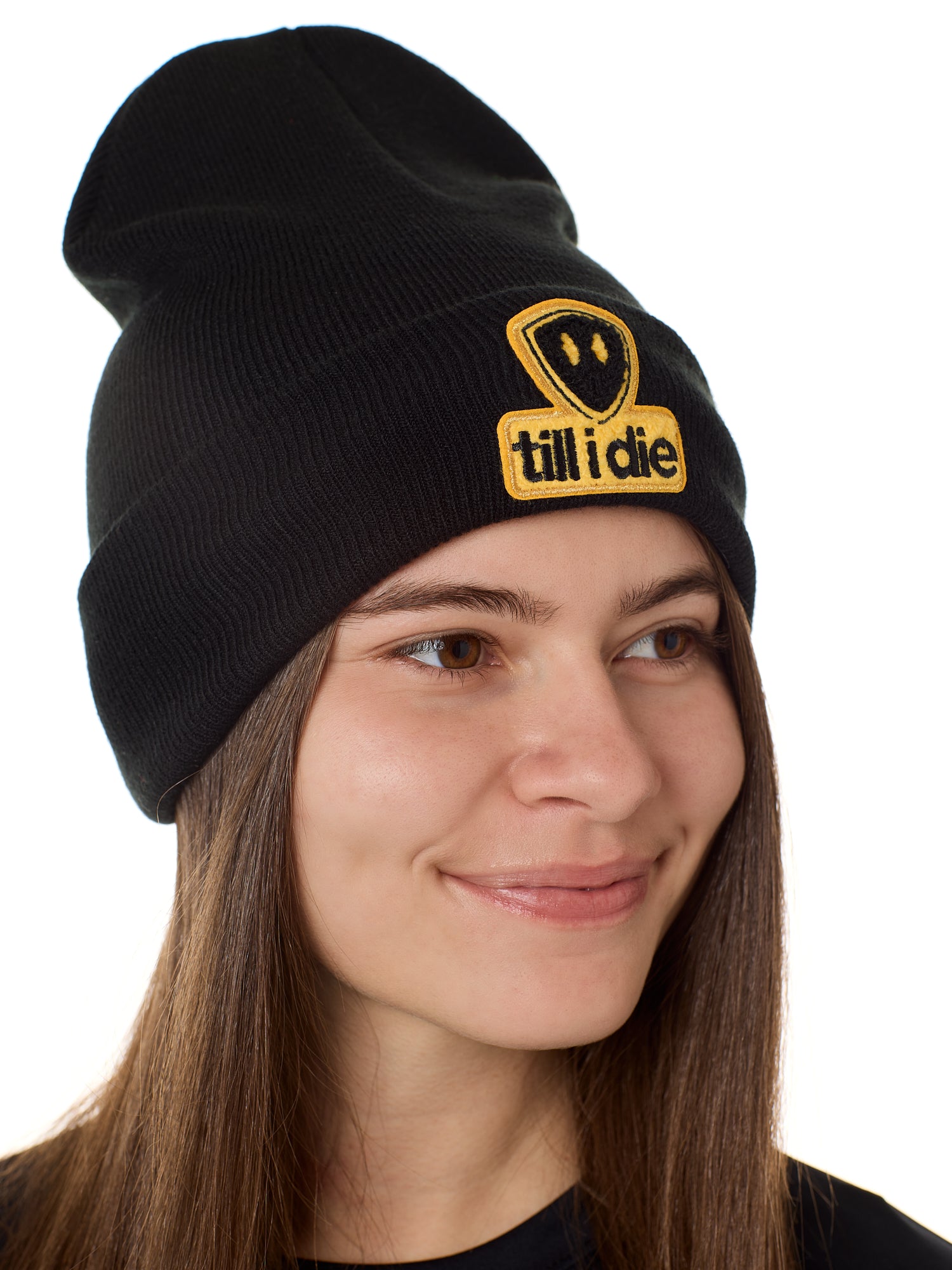 Sideview of a female model wearing a black knit beanie, featuring a cuff and a chenille patch of the Till I Die logo in yellow and black.