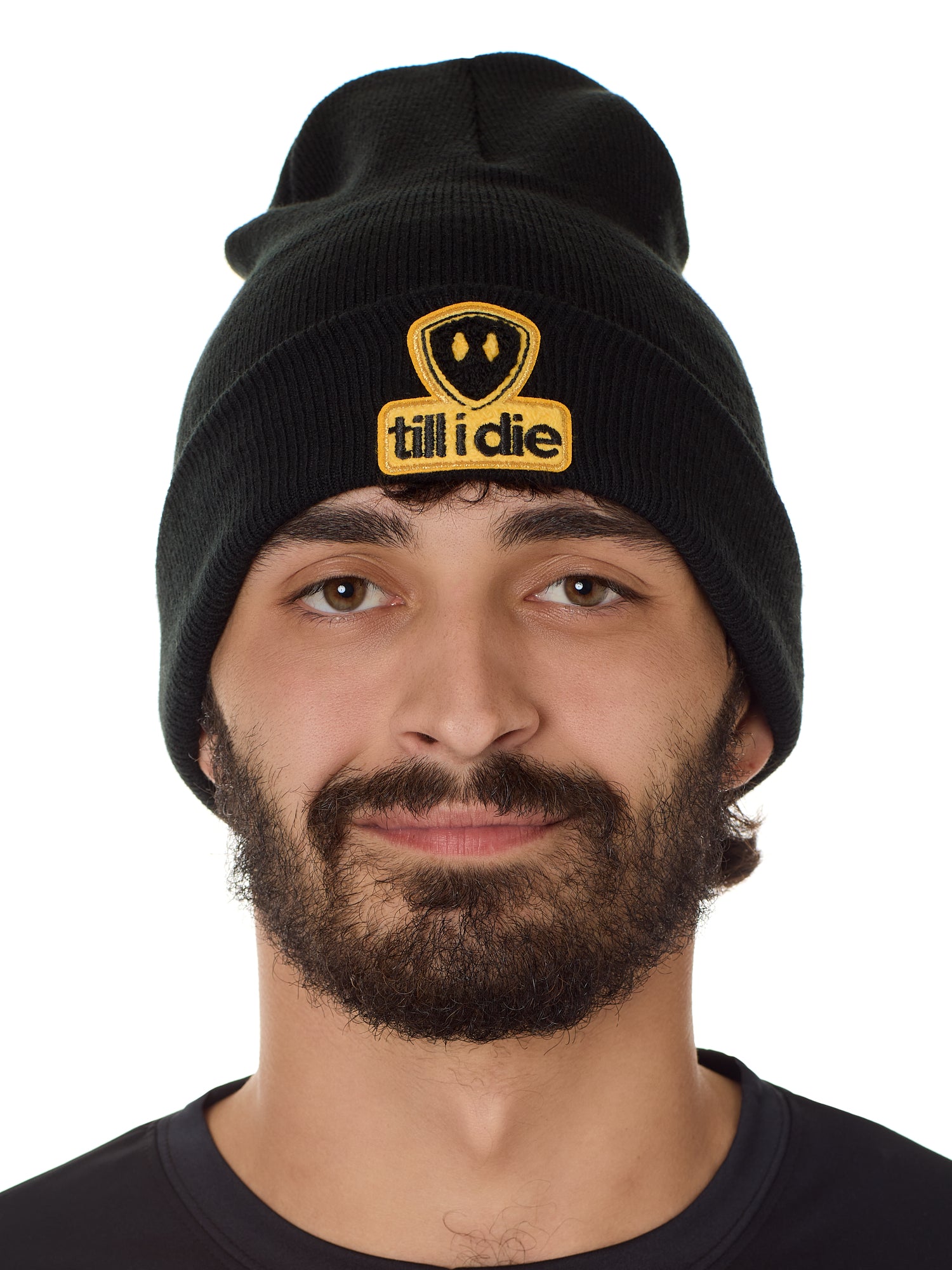 Front view of a male model wearing a black knit beanie, featuring a cuff and a chenille patch of the Till I Die logo in yellow and black.