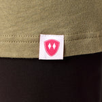 Close-up detail of a pink Double Diamond Shield woven hem label on a Light Olive Green tee shirt.