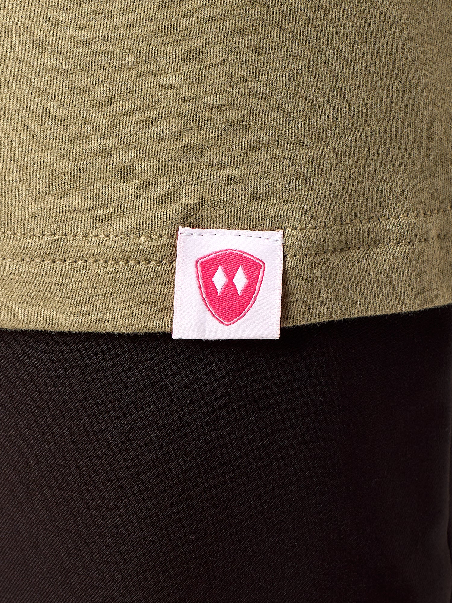 Close-up detail of a pink Double Diamond Shield woven hem label on a Light Olive Green tee shirt.