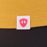 Close-up detail of a pink Double Diamond Shield woven hem label on a mustard tee shirt.