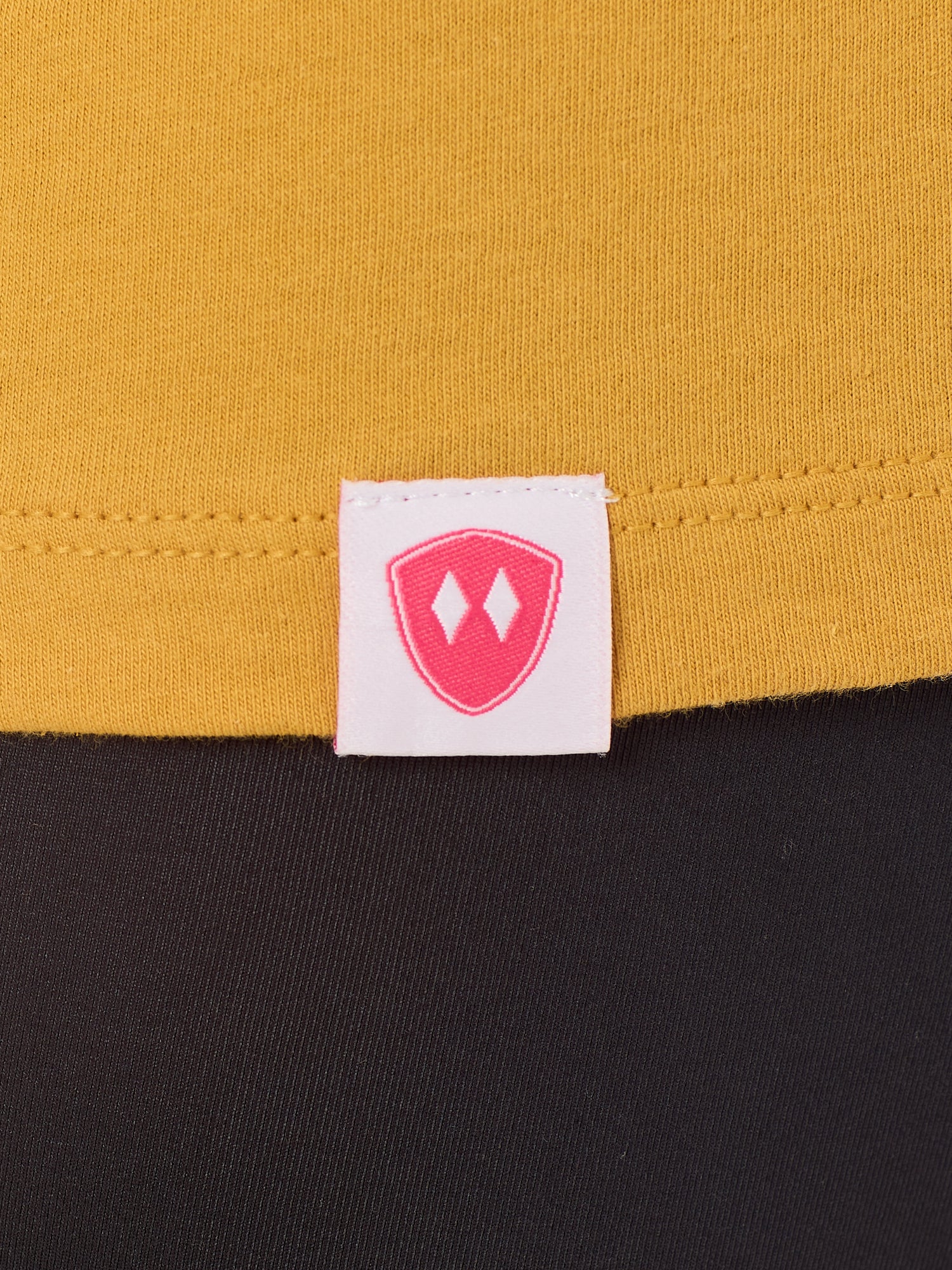 Close-up detail of a pink Double Diamond Shield woven hem label on a mustard tee shirt.