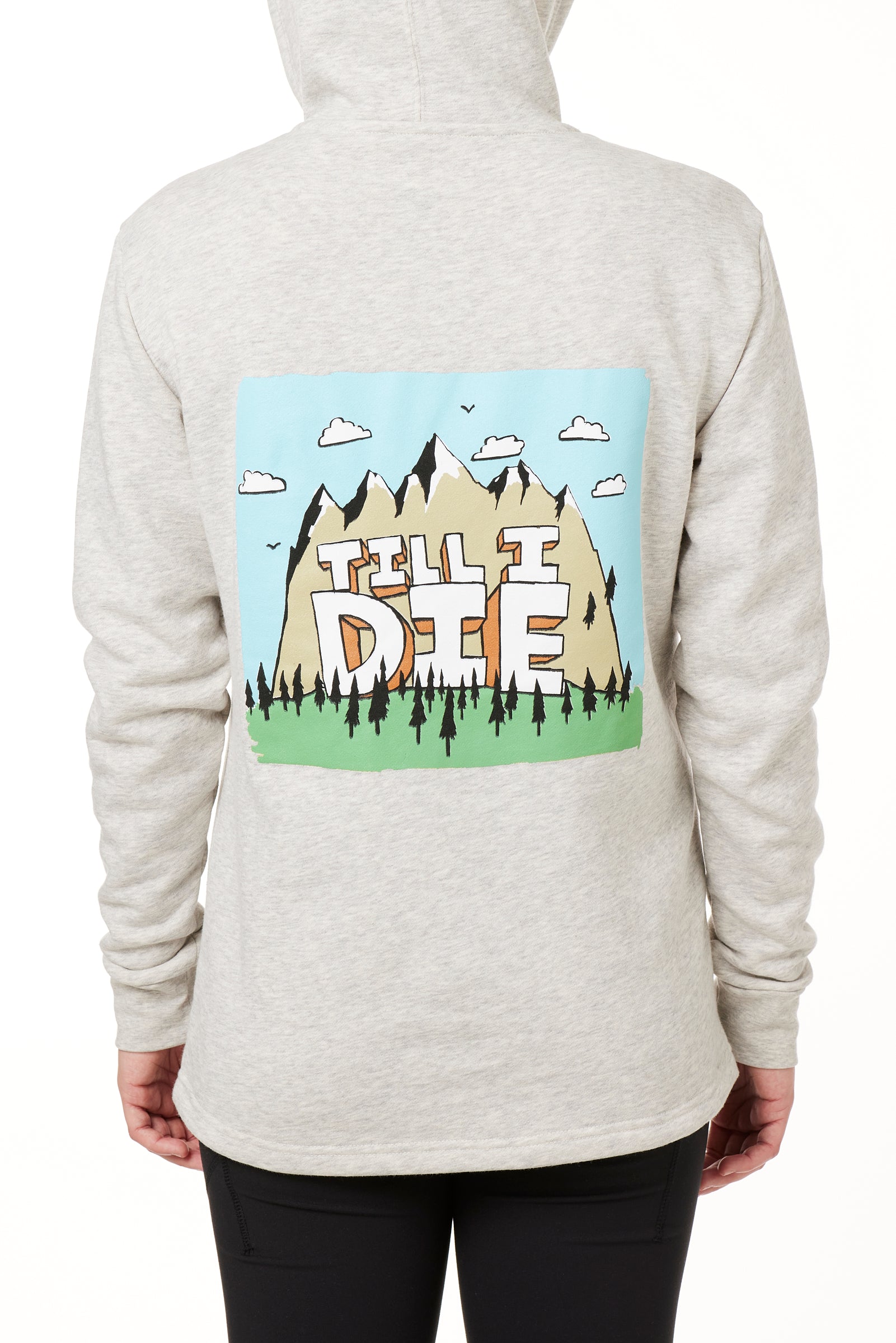 FEMALE MODEL WEARING A HEATHER OATMEAL HOODIE WITH HOOD UP, FEATURING A GRAPHIC OF A CARTOON MOUNTAIN THAT HAS TILL I DIE ACROSS IT