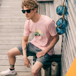 Male model in a beach town setting, sitting on the boardwalk wearing the surf truck tee and smoke and mirrors sunglasses