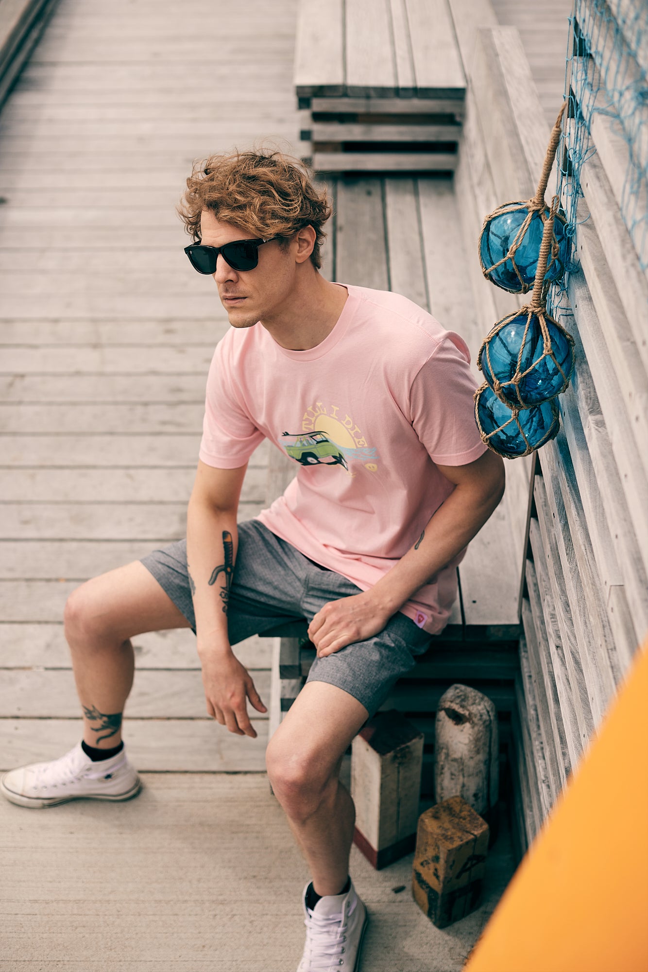 Male model in a beach town setting, sitting on the boardwalk wearing the surf truck tee and smoke and mirrors sunglasses