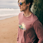 Male model leaning against an oceanside pier wearing the Surf Truck long sleeve tee and Electric Jungle sunglasses. The setting evokes laid-back coastal vibes with a bold, surf-inspired look.