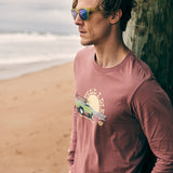 Male model leaning against an oceanside pier wearing the Surf Truck long sleeve tee and Electric Jungle sunglasses. The setting evokes laid-back coastal vibes with a bold, surf-inspired look.