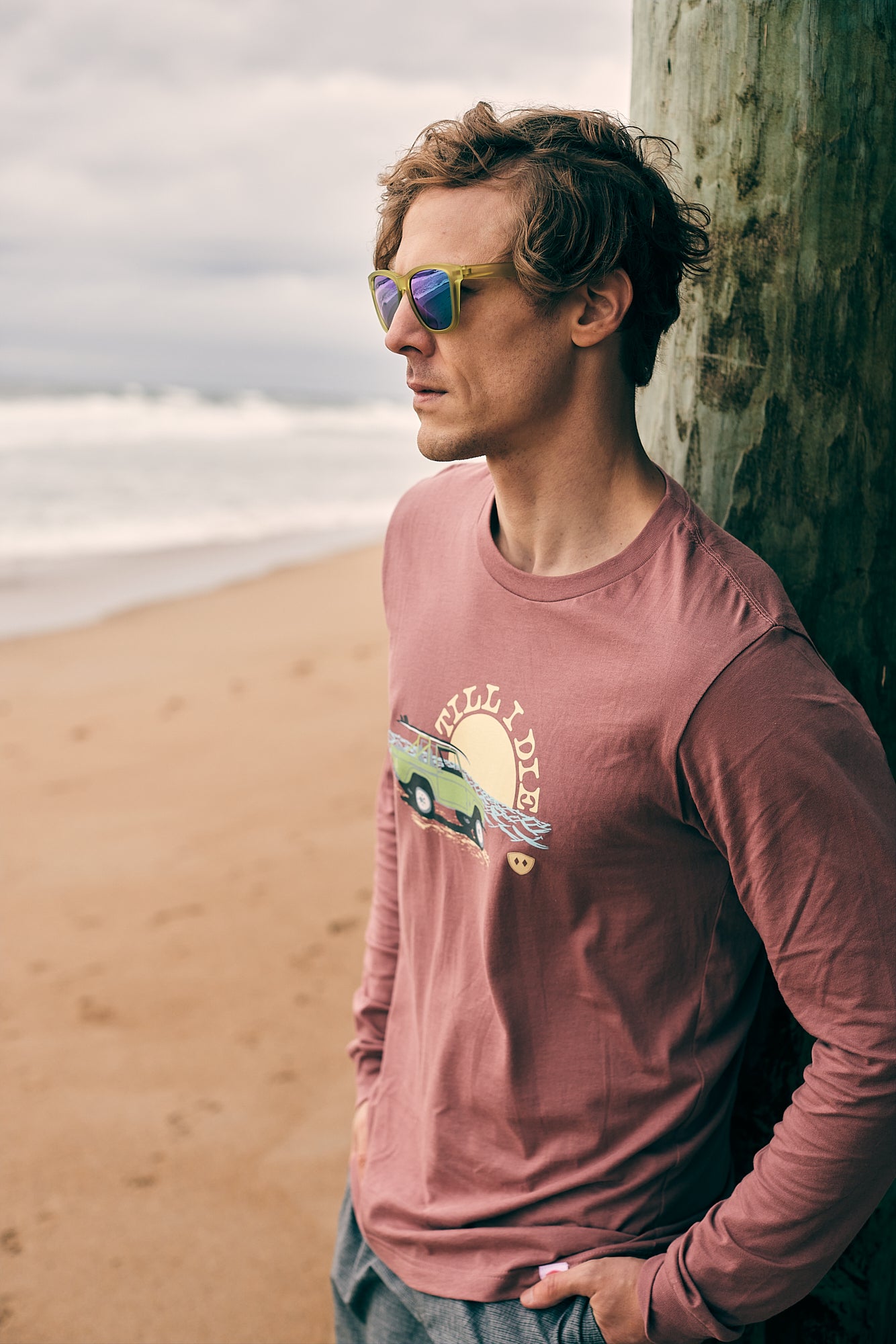 Male model leaning against an oceanside pier wearing the Surf Truck long sleeve tee and Electric Jungle sunglasses. The setting evokes laid-back coastal vibes with a bold, surf-inspired look.