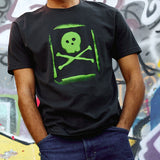 Man wearing the Graffiti Tee in Graphite Black, standing in front of a spray paint mural. The tee features a lime green spray-painted skull and crossbones graphic with a stencil-style overspray effect.