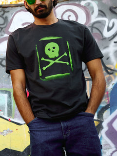 Man wearing the Graffiti Tee in Graphite Black, standing in front of a spray paint mural. The tee features a lime green spray-painted skull and crossbones graphic with a stencil-style overspray effect.