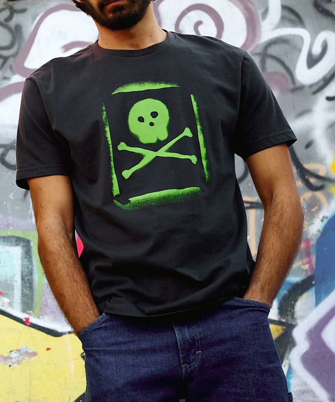 Man wearing the Graffiti Tee in Graphite Black, standing in front of a spray paint mural. The tee features a lime green spray-painted skull and crossbones graphic with a stencil-style overspray effect.