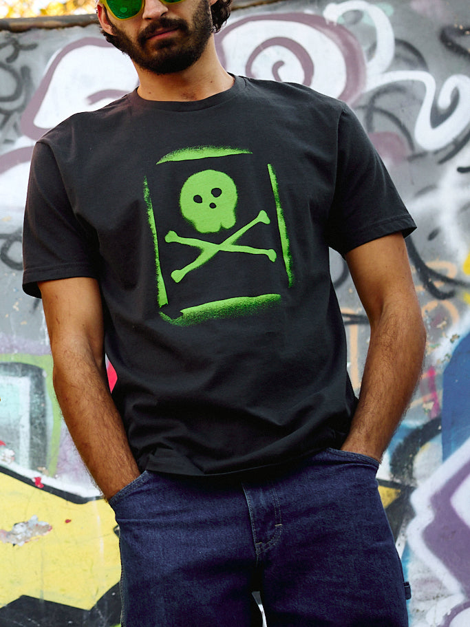 Man wearing the Graffiti Tee in Graphite Black, standing in front of a spray paint mural. The tee features a lime green spray-painted skull and crossbones graphic with a stencil-style overspray effect.