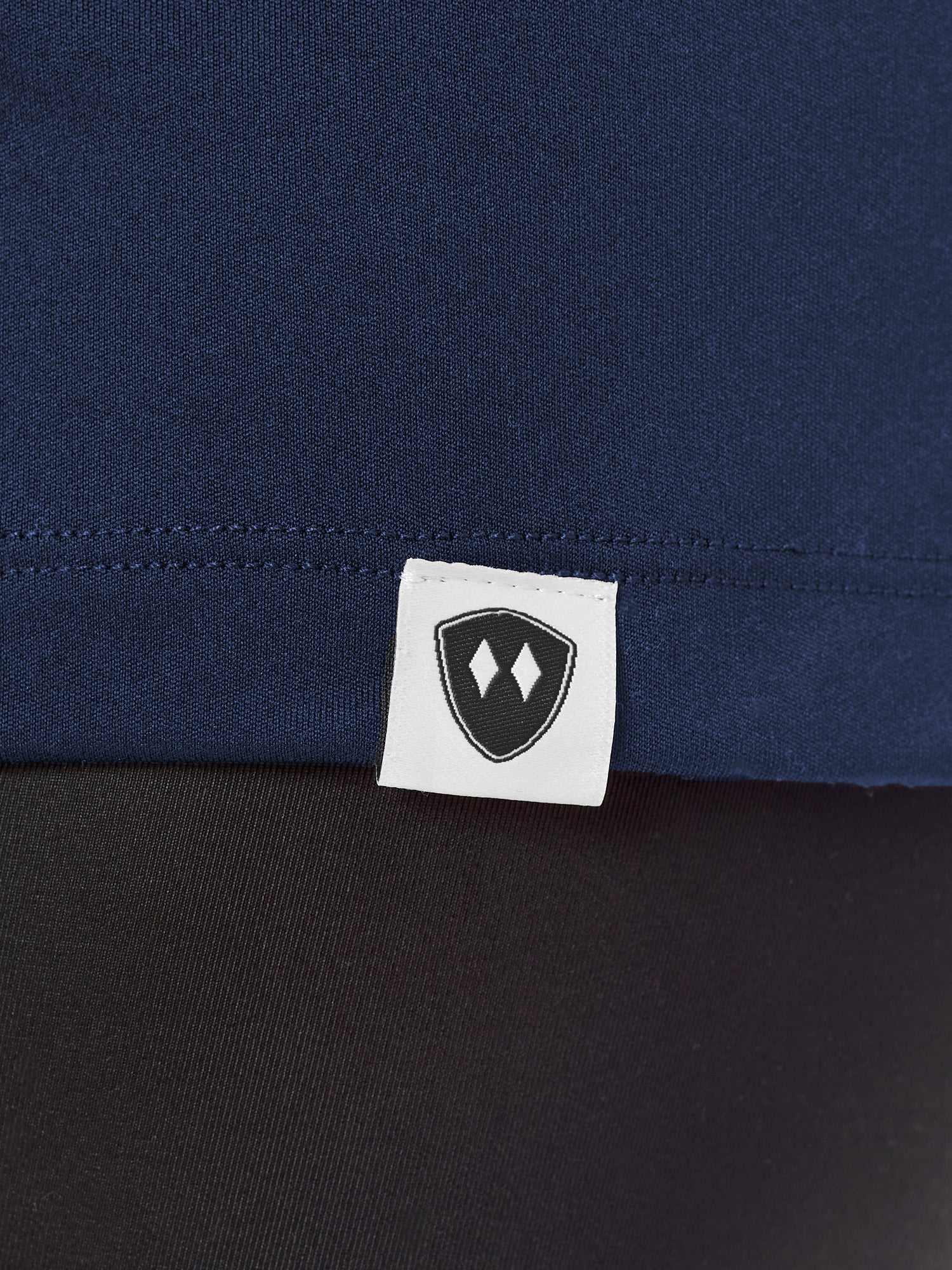 Close-up detail of a black Double Diamond Shield woven hem label on a navy performance long sleeve shirt.