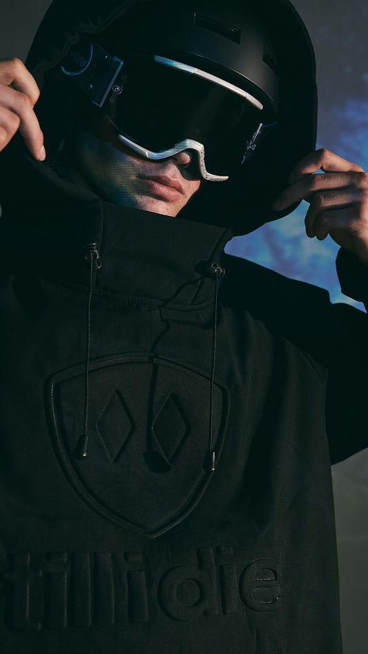 Male model wearing goggles and the black Performance Pullover Jacket with the hood up, gripping the hood with both hands. The moody scene features a snowy landscape projection in the background, with lighting that highlights the embossed Till I Die logo on the front of the jacket.