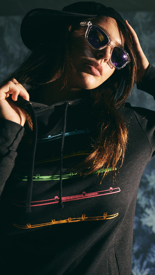 Female model wearing the Shot Ski Hoodie, Violet Haze sunglasses, and a sideways black trucker hat. She is gripping the collar with one hand and adjusting the hat with the other. The scene features a moody vibe with a snowy landscape projection in the background.