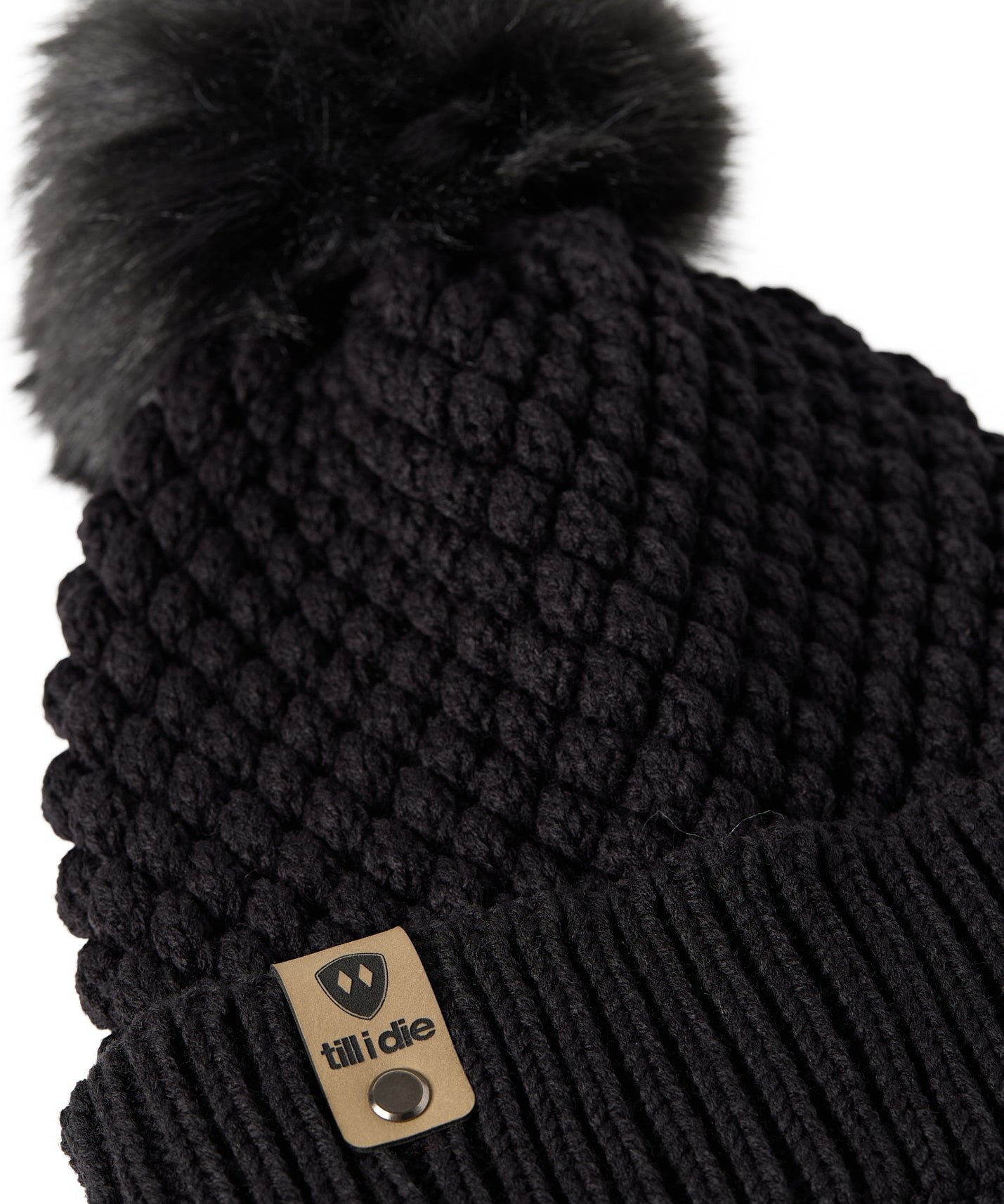 Close-up flat lay of the Diamond Knit Pom Beanie showing the textured diamond knit pattern, faux fur pom, and logo leather tab.