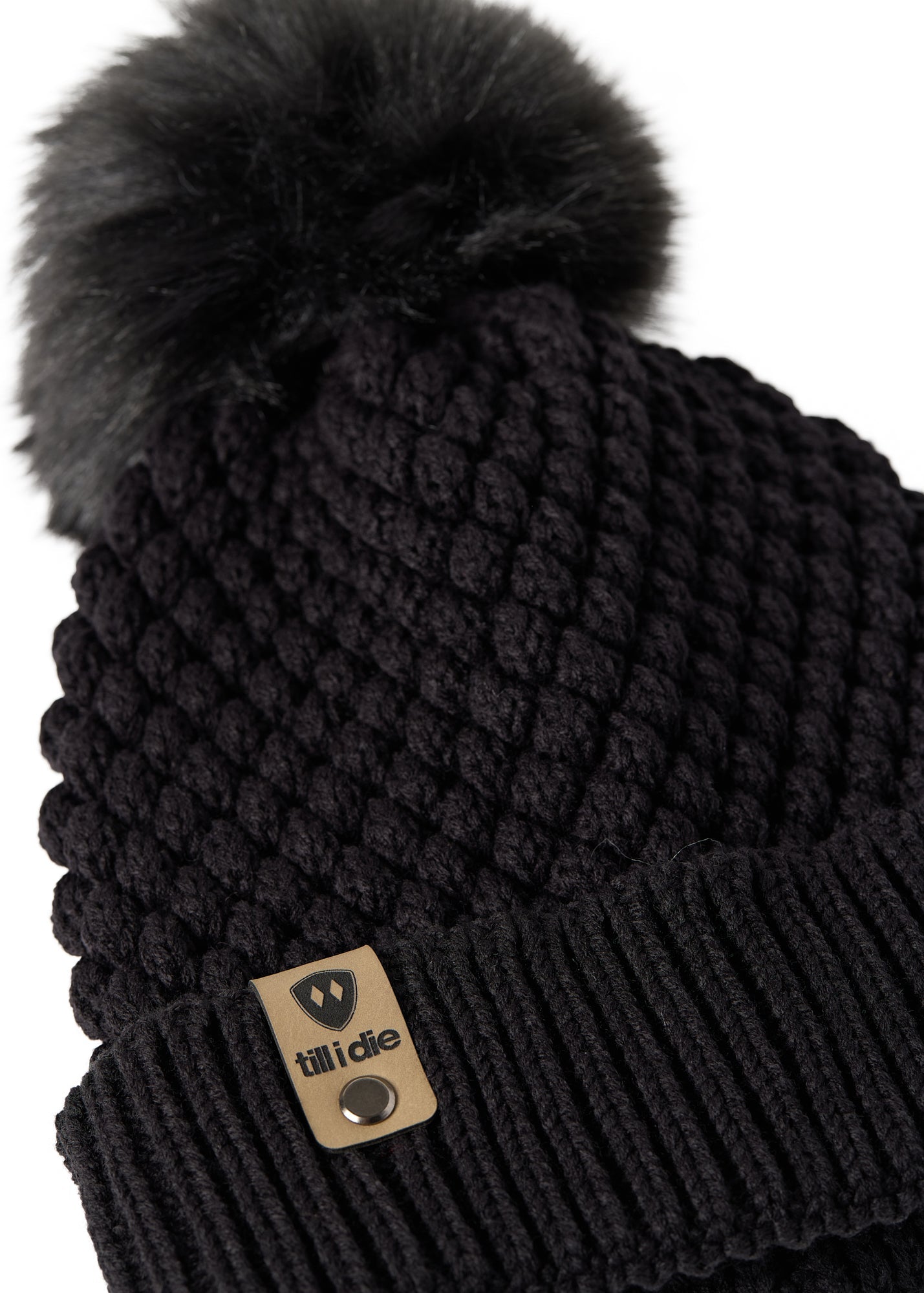 Close-up flat lay of the Diamond Knit Pom Beanie showing the textured diamond knit pattern, faux fur pom, and logo leather tab.