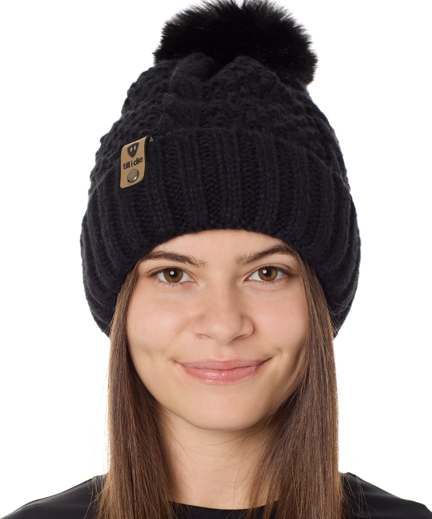 Front view of a female model wearing the black knit pom beanie hat in black on a white background. the hat features a matching pom pom, leather tab with embossed black till i die logo and nickel fastener. 