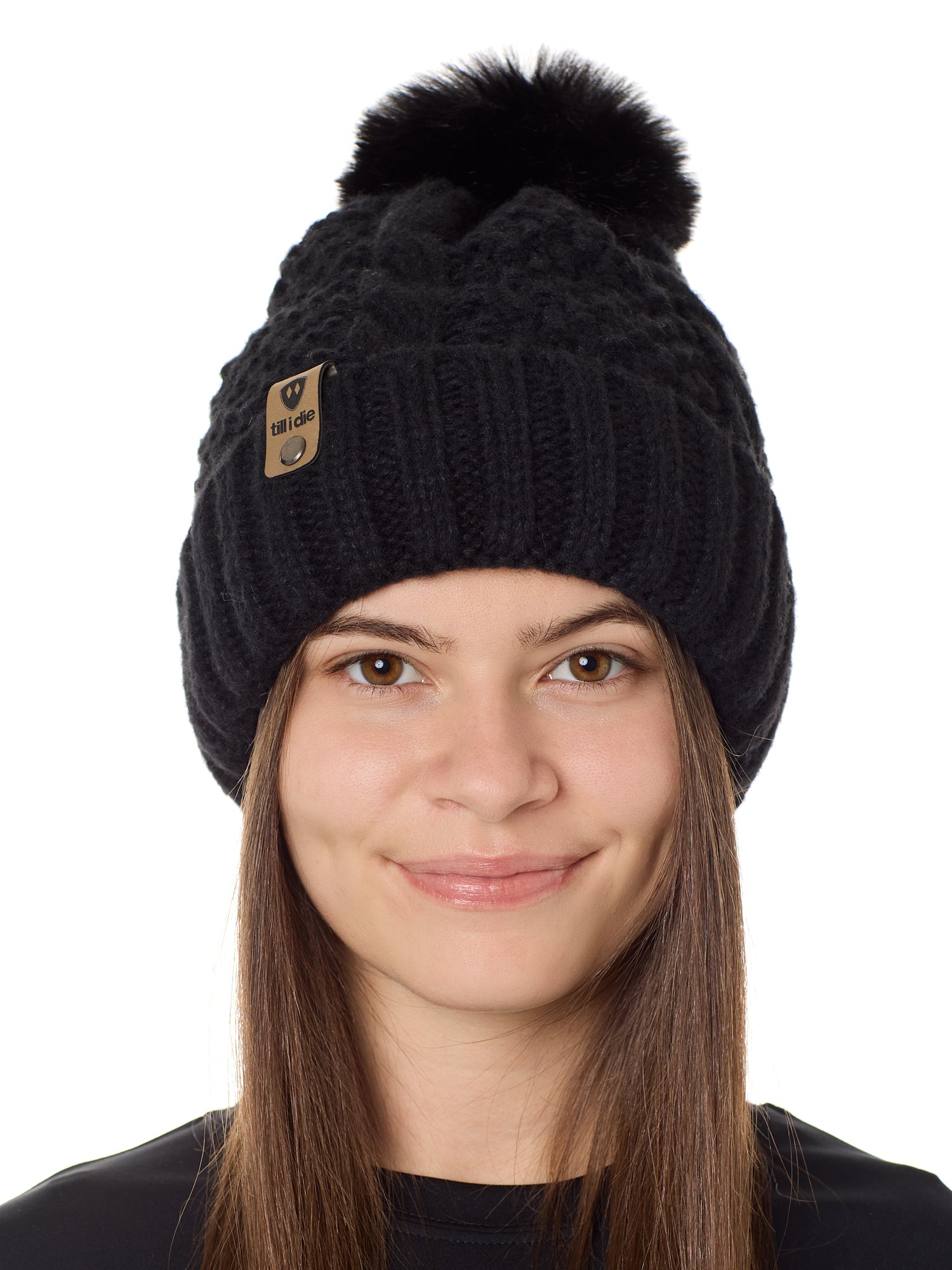 Front view of a female model wearing the black knit pom beanie hat in black on a white background. the hat features a matching pom pom, leather tab with embossed black till i die logo and nickel fastener. 