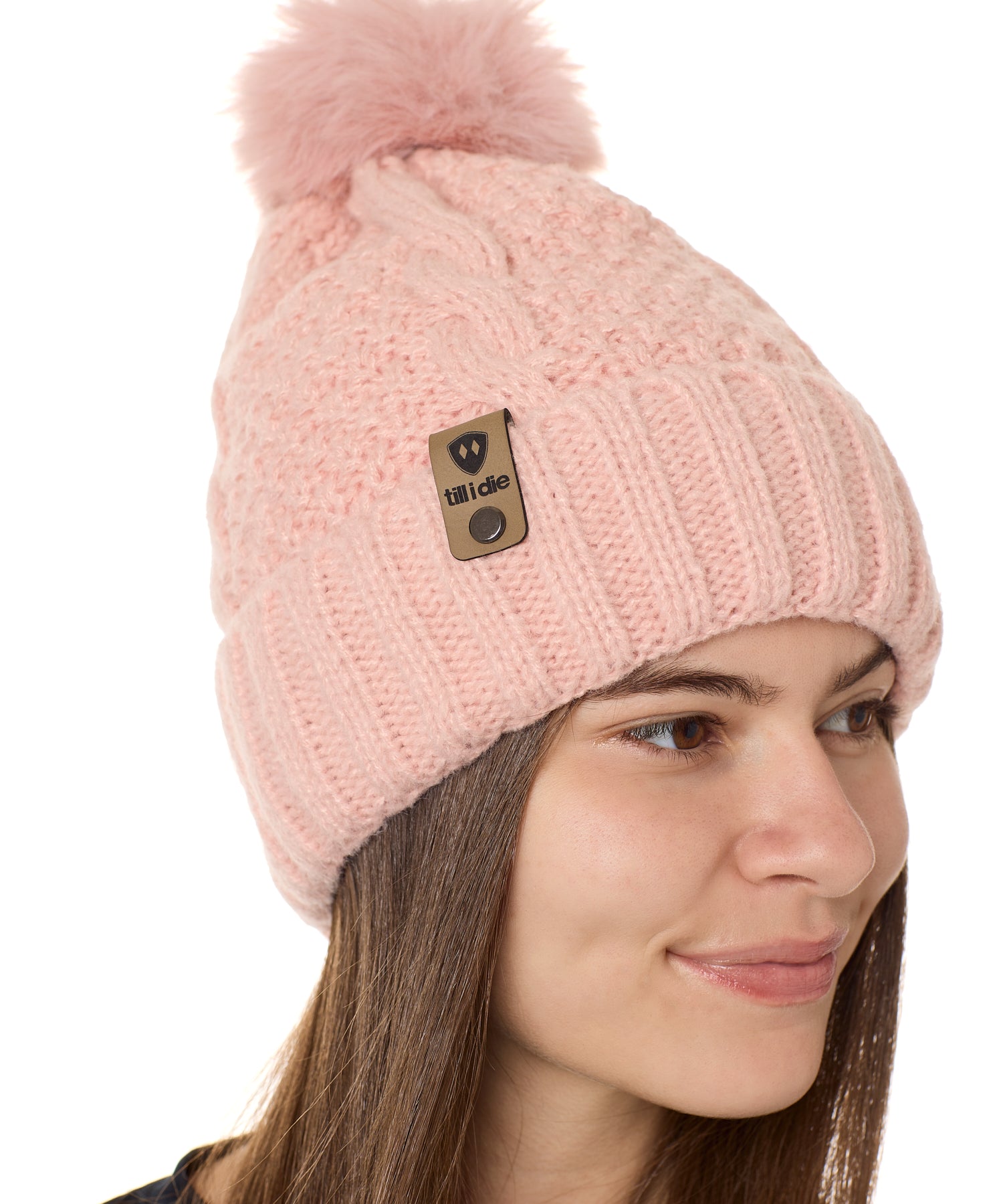 side view of a female model wearing a blush knit pom beanie featuring a matching pom pom, cuff and leather till i die logo tab with a nickel fastener.