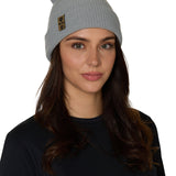 Female model wearing the Till I Die Leather Tab Knit Beanie in cadet blue, styled with the cuff folded to show the branded leather logo tab