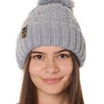 front view of a female model wearing a gray knit pom beanie featuring a matching pom pom, cuff and leather till i die logo tab with a nickel fastener.