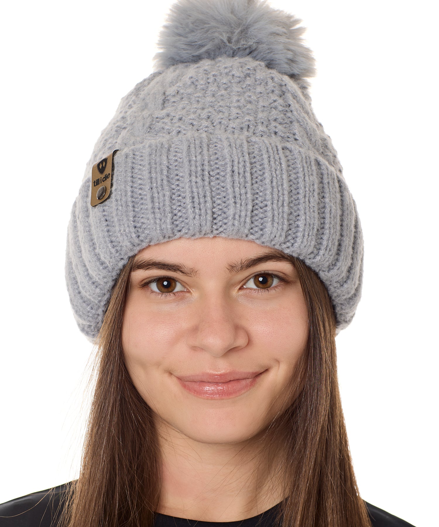 front view of a female model wearing a gray knit pom beanie featuring a matching pom pom, cuff and leather till i die logo tab with a nickel fastener.