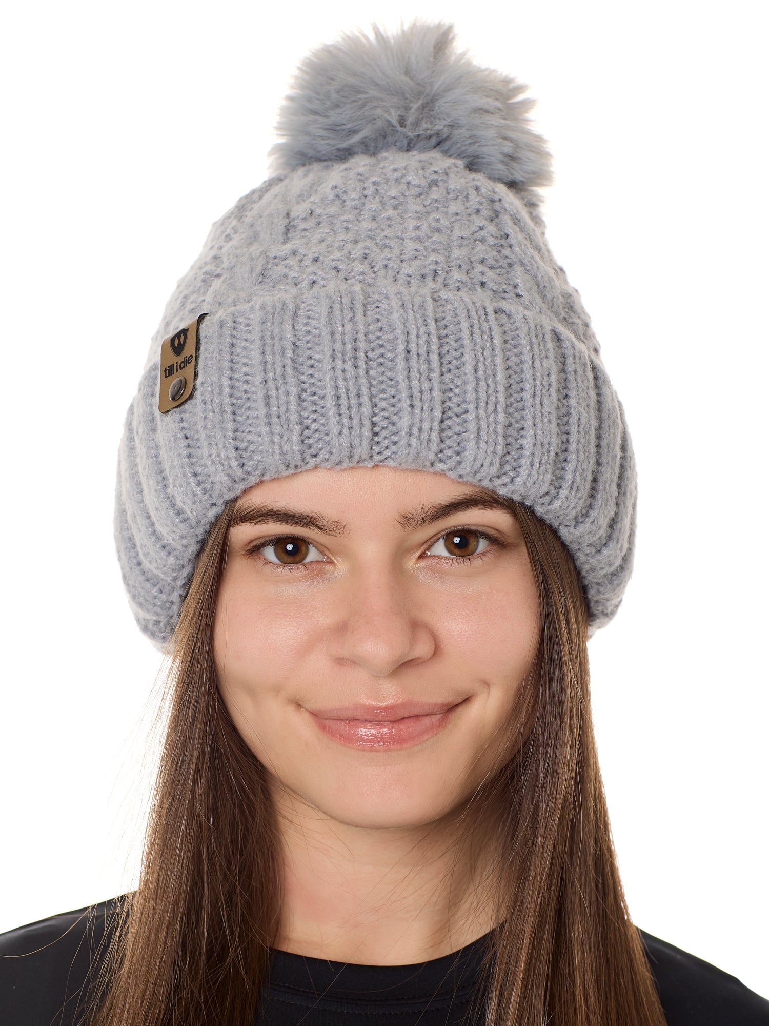 front view of a female model wearing a gray knit pom beanie featuring a matching pom pom, cuff and leather till i die logo tab with a nickel fastener.