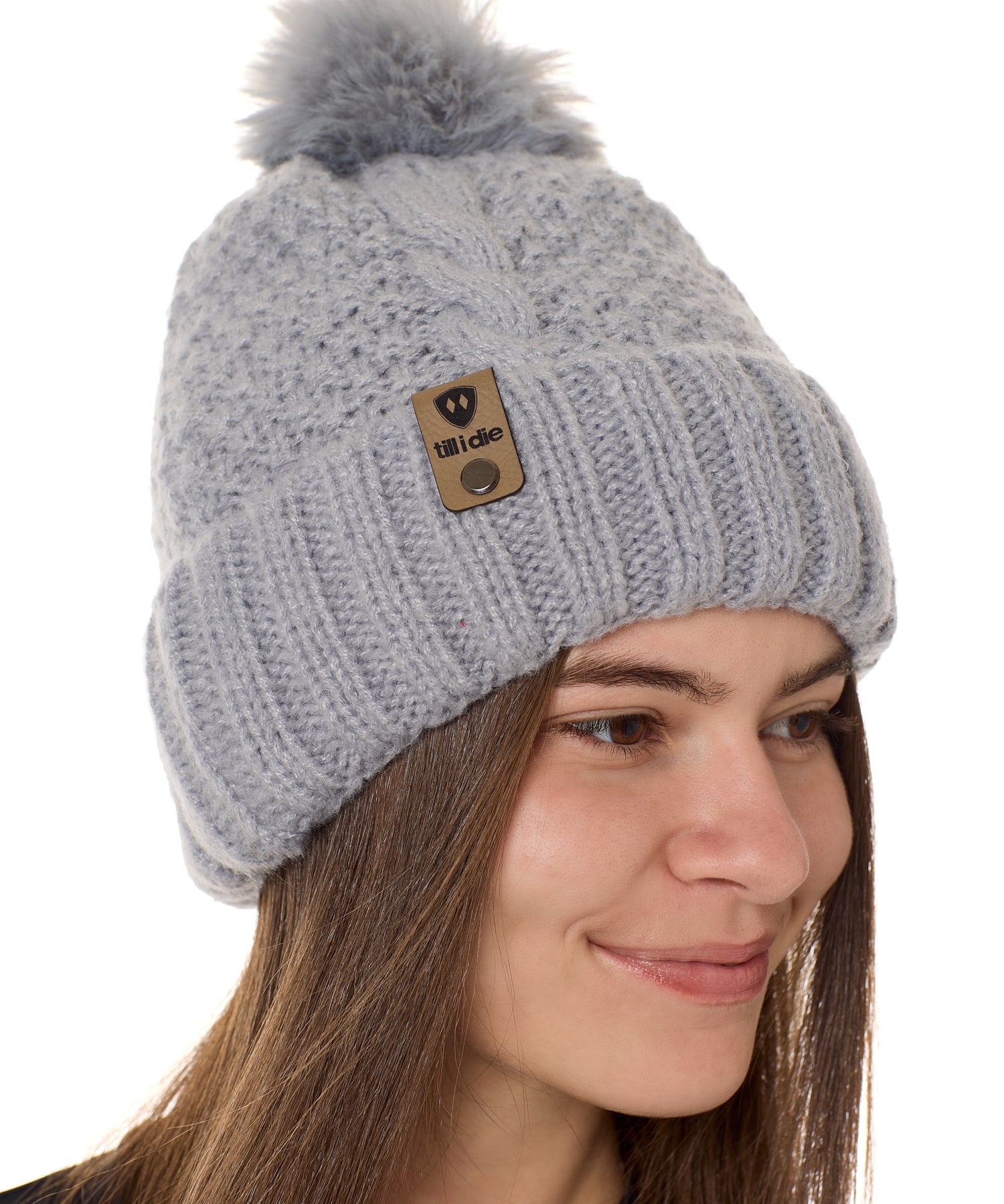 side view of a female model wearing a gray knit pom beanie featuring a matching pom pom, cuff and leather till i die logo tab with a nickel fastener.